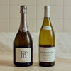 Two wine bottles standing side by side on a light stone surface against a beige tiled wall. The left bottle is a dark sparkling wine with a silver foil neck and a white label reading 'bis' and 'Bellemonde'; the right bottle is a pale green Chablis white wine with a white label reading 'Chablis 1er Cru' and 'Fourchaumie'.