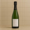 A bottle of Hure Freres Invitation Champagne, featuring a dark green glass bottle, a black foil top, and a white label, set against a beige tiled background.