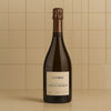 A bottle of L'Assemblage Champagne Pertois-Morlet standing on a light beige surface against a beige tiled background.