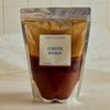 A stand-up pouch of Portside Lobster Bisque on a light marble countertop with a tiled backsplash. The pouch is made of reflective silver foil, showing a layered sauce inside with a white label in the center that reads 'LOBSTER BISQUE' in blue text.