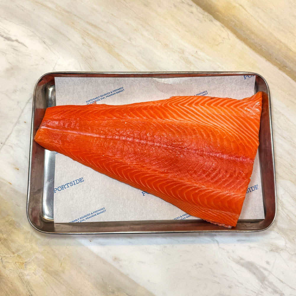 A raw salmon fillet with vibrant orange-pink flesh lies on parchment paper inside a rectangular metal baking tray, set on a light beige marble surface.