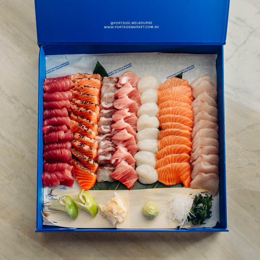 A large sashimi and sushi gift box from Portside Market, displayed open on a light marble surface. The blue box is filled with neatly arranged raw fish slices including tuna, salmon, mackerel, sea urchin, and white fish, along with garnishes like wasabi, pickled ginger, and lime.