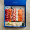 A large sashimi and sushi gift box from Portside Market, displayed open on a light marble surface. The blue box is filled with neatly arranged raw fish slices including tuna, salmon, mackerel, sea urchin, and white fish, along with garnishes like wasabi, pickled ginger, and lime.