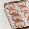 A metal baking tray lined with Portiste parchment paper, holding several scallop shells arranged in a circular pattern, each with a pale beige pearl or custard-like center.
