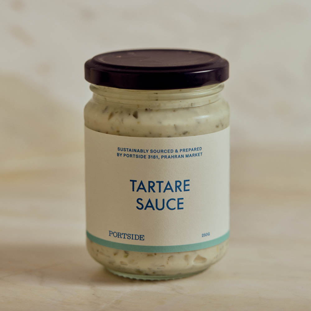 A glass jar of Portside Tartare Sauce with a cream-colored label and a dark purple lid, set against a neutral, light beige background.