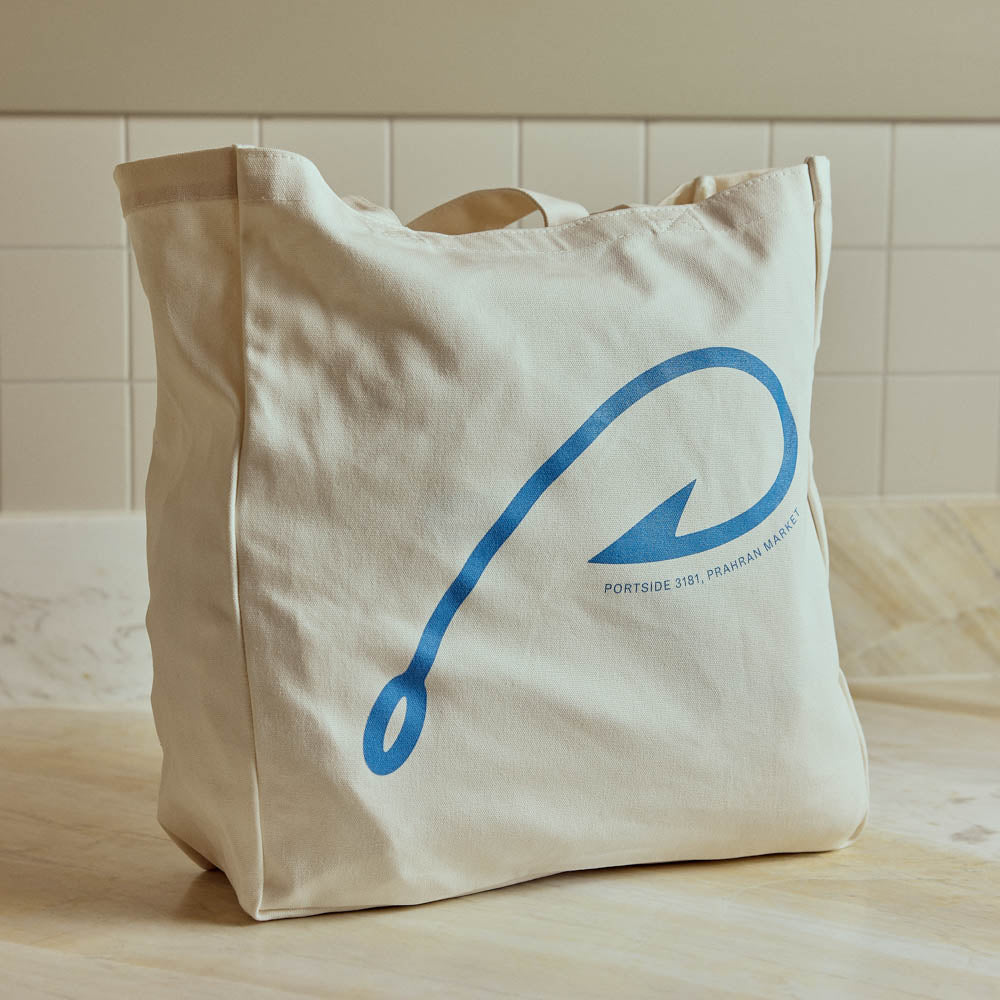 A natural canvas tote bag with a blue graphic of a hook and the text 'PORTSIDE 3RD TRINITY MARKET' printed on the front, set on a light wooden surface against a tiled wall.