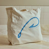 A natural canvas tote bag with a blue graphic of a hook and the text 'PORTSIDE 3RD TRINITY MARKET' printed on the front, set on a light wooden surface against a tiled wall.