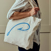 A person wearing a light-colored shirt carries a cream canvas tote bag over their shoulder. The bag features a blue graphic of a hook with a fish inside and the text 'POSTSIDE 205, PRAGUAN MARKET' printed below.