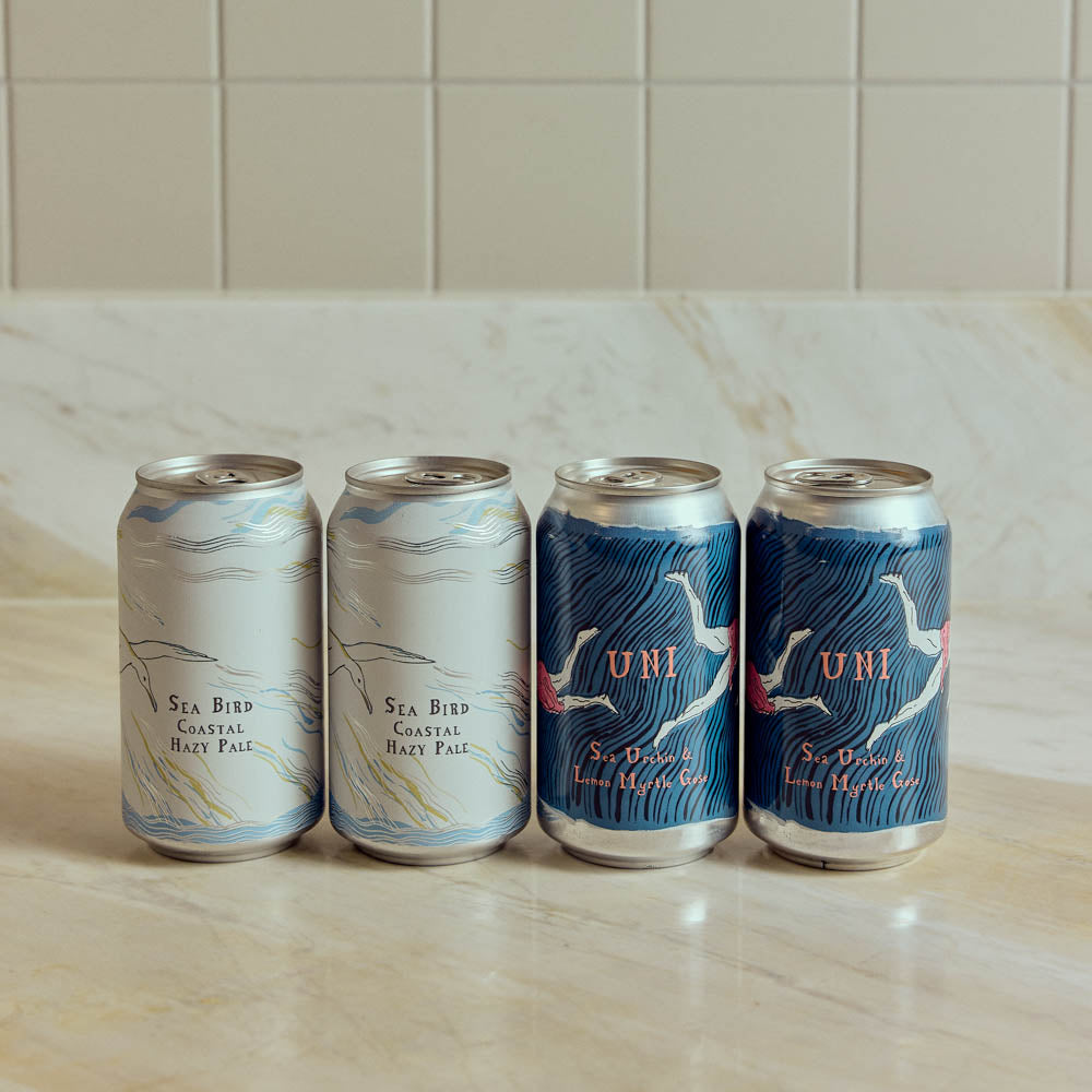 Four aluminum craft beer cans lined up on a light marble countertop against a beige tiled backsplash. Two cans are silver with pale blue and white wave-like illustrations and the text 'Sea Bird Coastal Hazy Pale,' and two cans are dark blue with white bird illustrations and the brand name 'UNI' in orange.