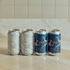 Four aluminum craft beer cans lined up on a light marble countertop against a beige tiled backsplash. Two cans are silver with pale blue and white wave-like illustrations and the text 'Sea Bird Coastal Hazy Pale,' and two cans are dark blue with white bird illustrations and the brand name 'UNI' in orange.