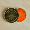 Yarra Valley Smoked Salmon Pearls in a round metal tin, shown open with the dark green branded lid on the left and bright orange salmon pearls visible on the right, set on a light marble surface.